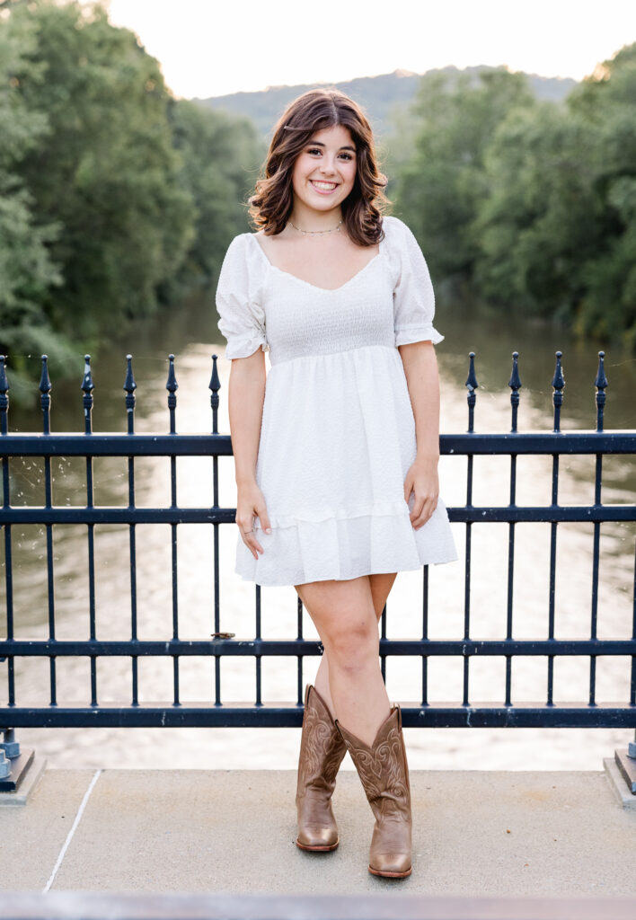 Girl wearing a white dress and cow boy boots standing on a bridge with a black railing 