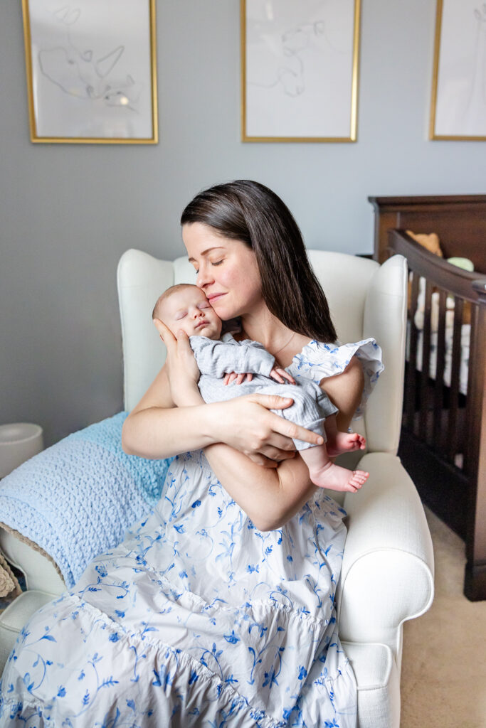 Mom resting cheek on newborns face during newborn photo session. Mom wearing neutral toned dress sitting on neutral chair. 