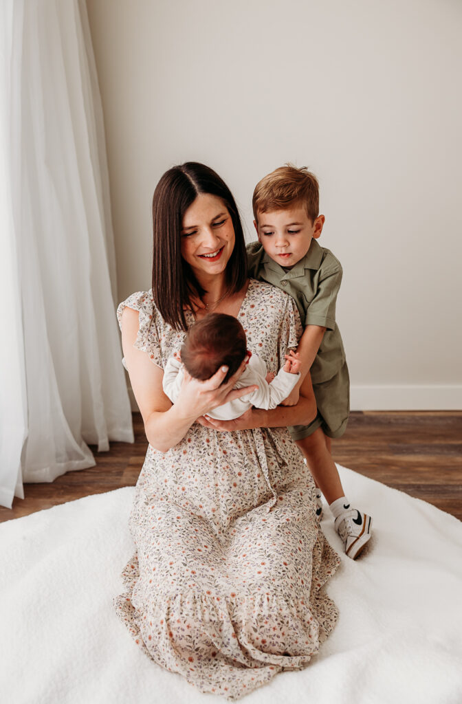 Mom holding newborn son with older brother looking at baby over moms shoulder
