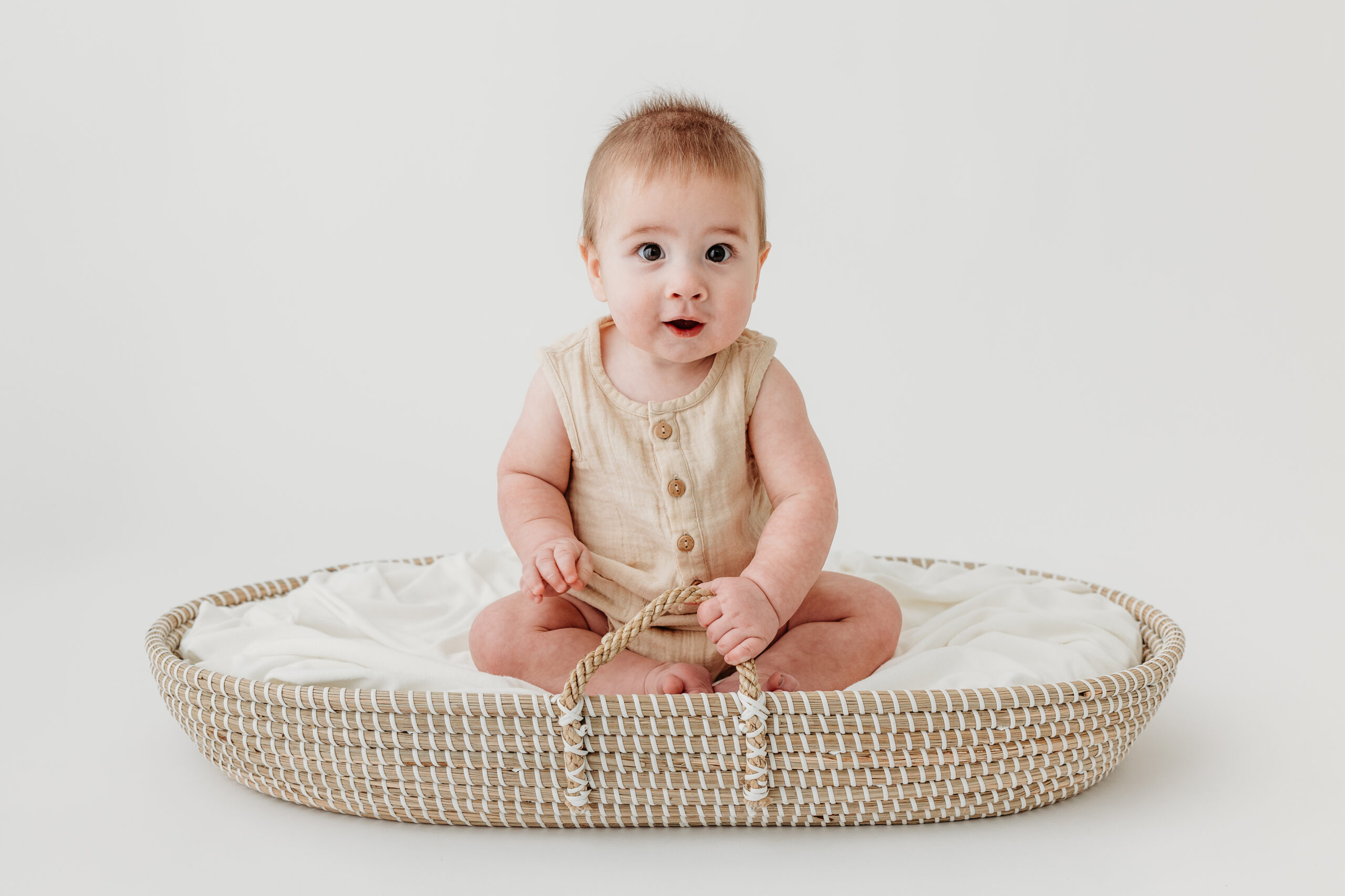A 6-month-old baby sits up independently in a basket, holding the handle, beaming with curiosity and joy during a milestone portrait session in Pittsburgh.
